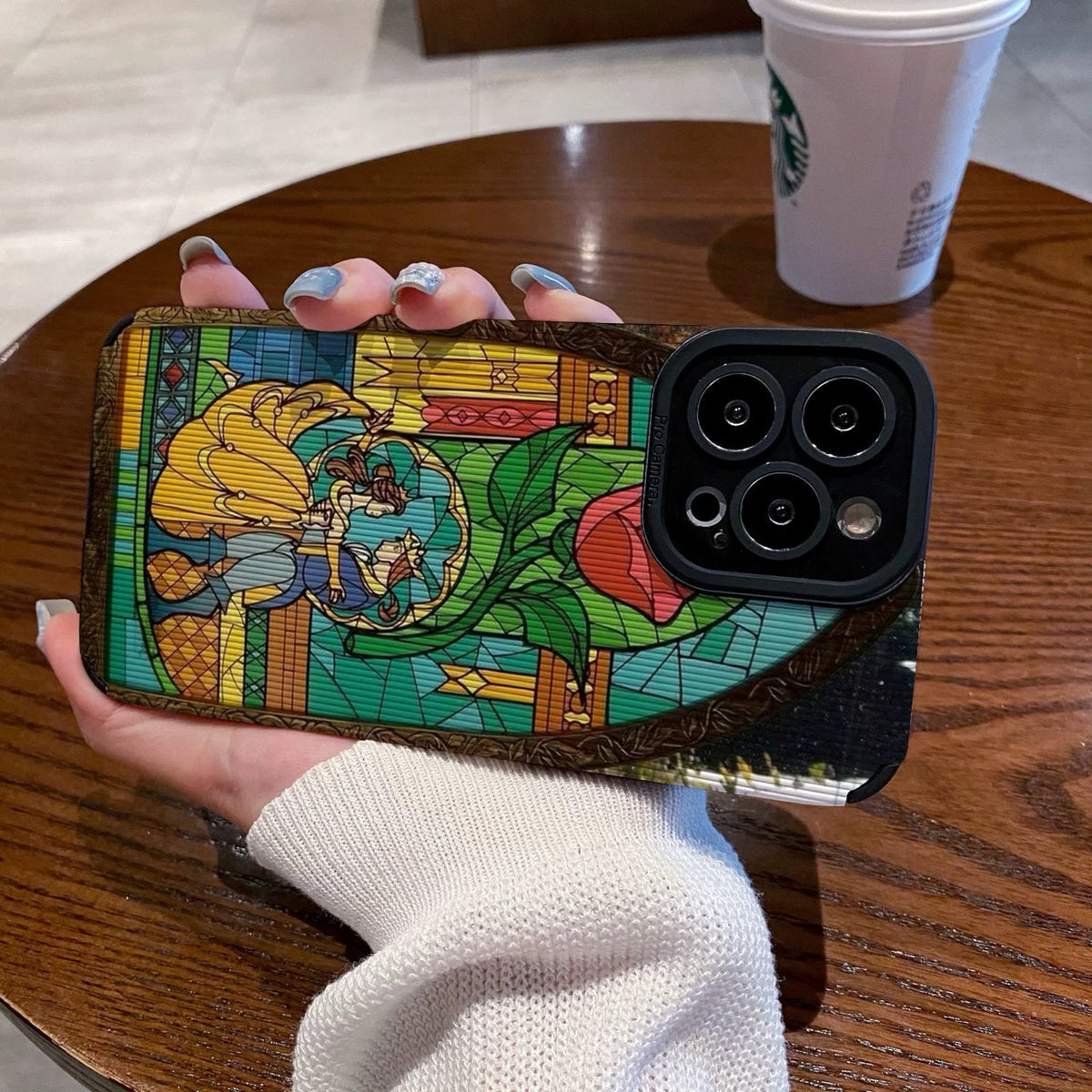 iPhone case with stained glass design with belle and the prince held by a hand on a wooden table with a Starbucks cup.