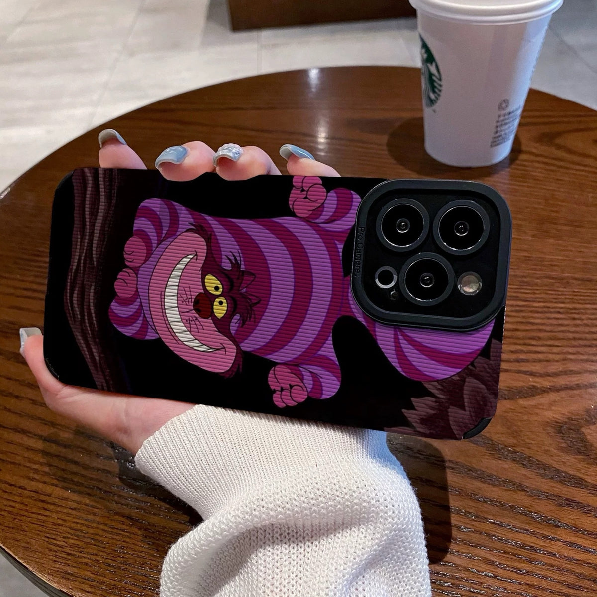 Hand holding a iPhone case with a Cheshire Cat of Alice in Wonderland on a wooden table with a Starbucks cup.