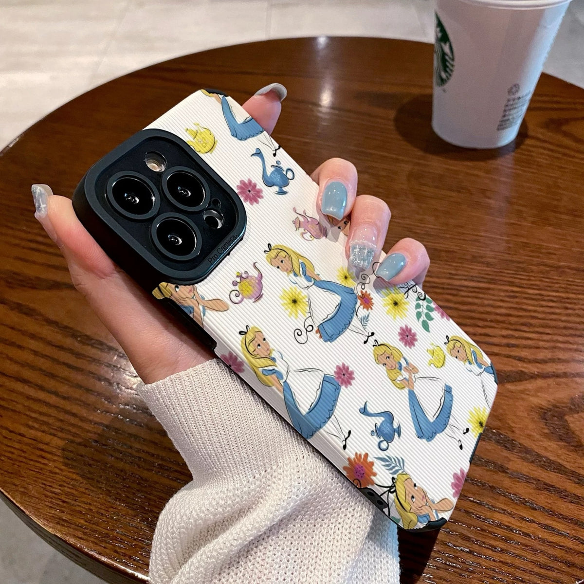 iPhone case with Alice in the Wonderland design held by a hand on a wooden table with a Starbucks cup in the background