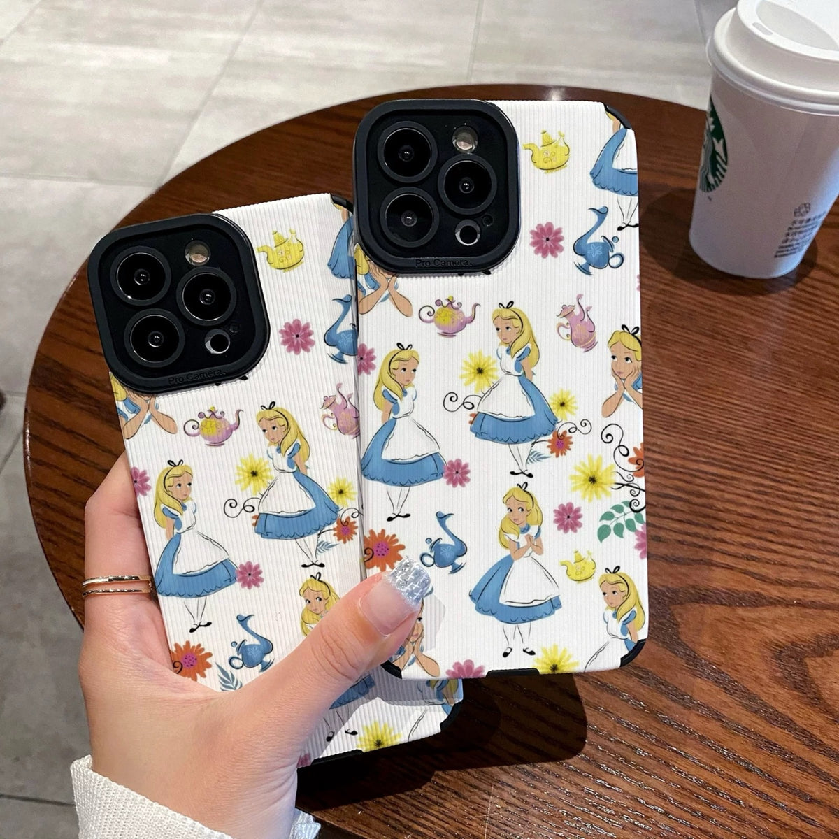 iPhone case with Alice in the Wonderland design held by a hand on a wooden table with a Starbucks cup in the background
