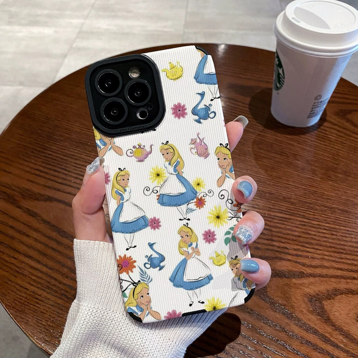 iPhone case with Alice in the Wonderland design held by a hand on a wooden table with a Starbucks cup in the background