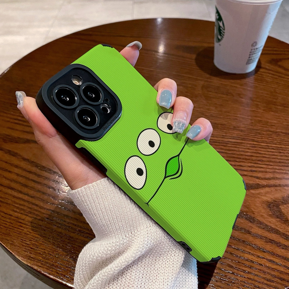 Person holding a green toy story alien phone case with cartoon face design on a wooden table with a Starbucks cup.