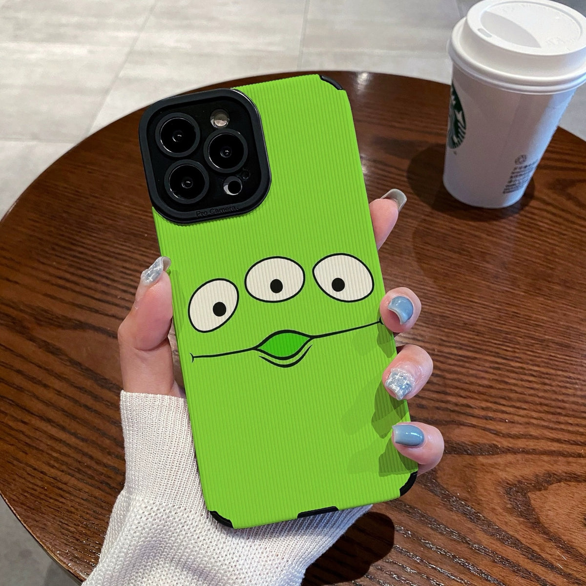 Person holding a green toy story alien phone case with cartoon face design on a wooden table with a Starbucks cup.