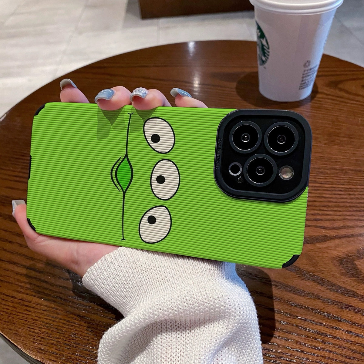 Person holding a green toy story alien phone case with cartoon face design on a wooden table with a Starbucks cup.