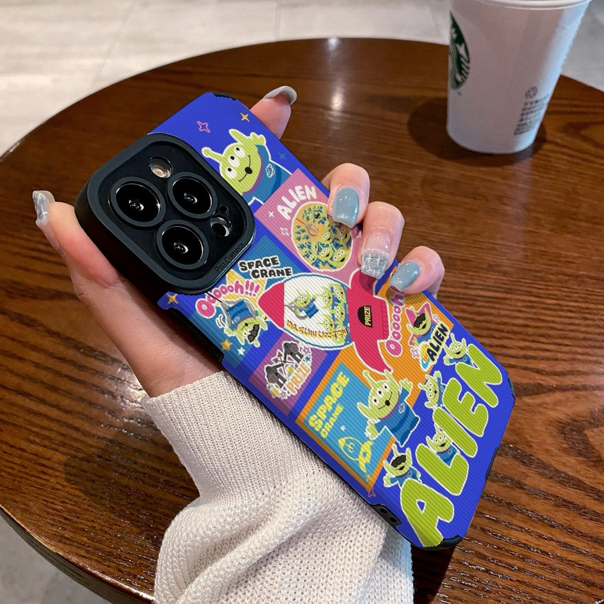 Colorful phone case with Toy Story alien design held by a person on a wooden table with a Starbucks cup in the background.