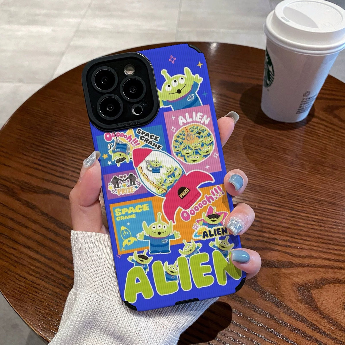 Colorful phone case with Toy Story alien design held by a person on a wooden table with a Starbucks cup in the background.
