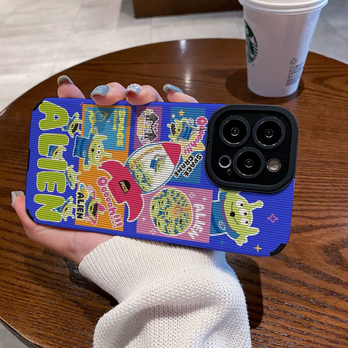 Colorful phone case with Toy Story alien design held by a person on a wooden table with a Starbucks cup in the background.