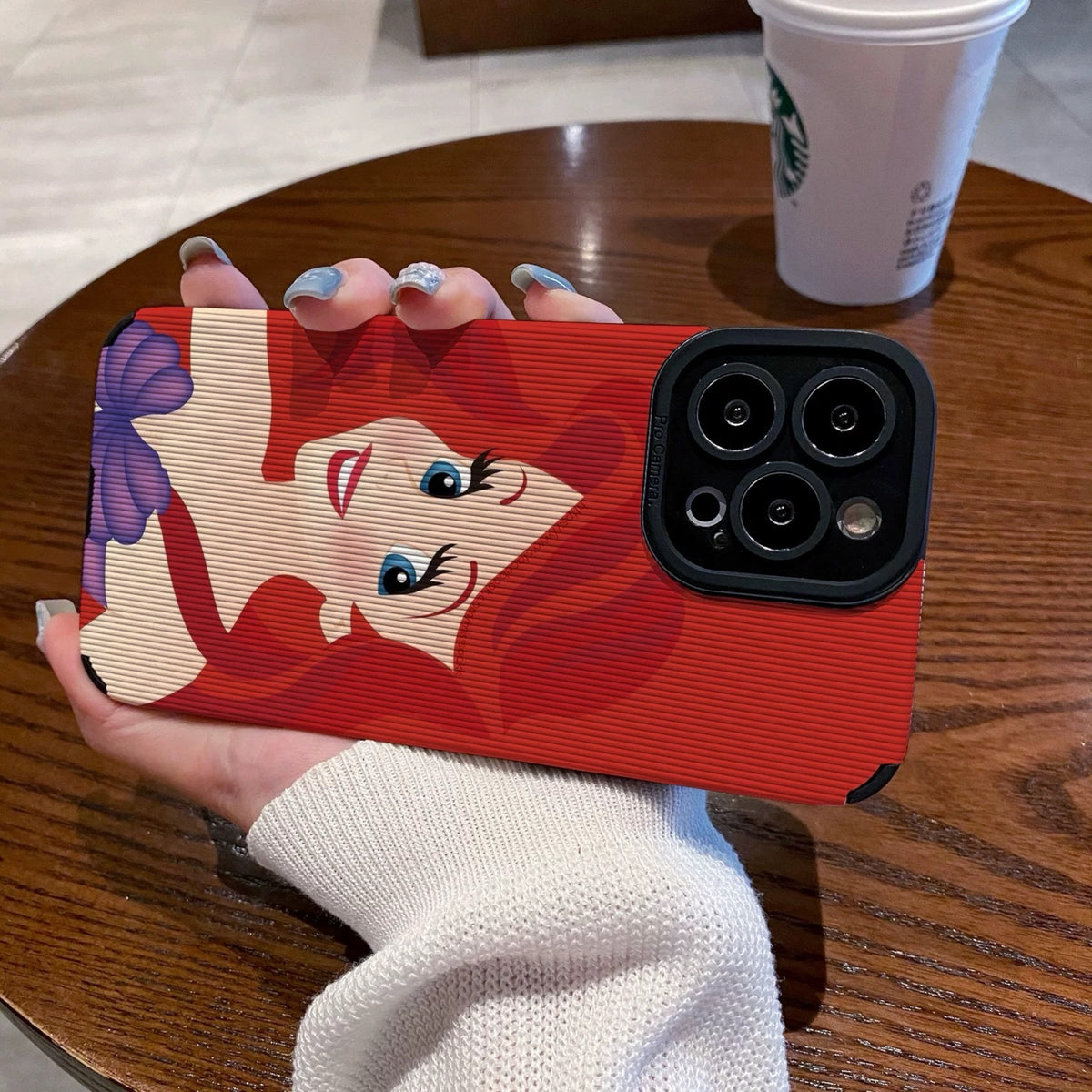 My Little Mermaid Ariel iPhone case with a character design held by a hand on a wooden table with a Starbucks cup in the background.
