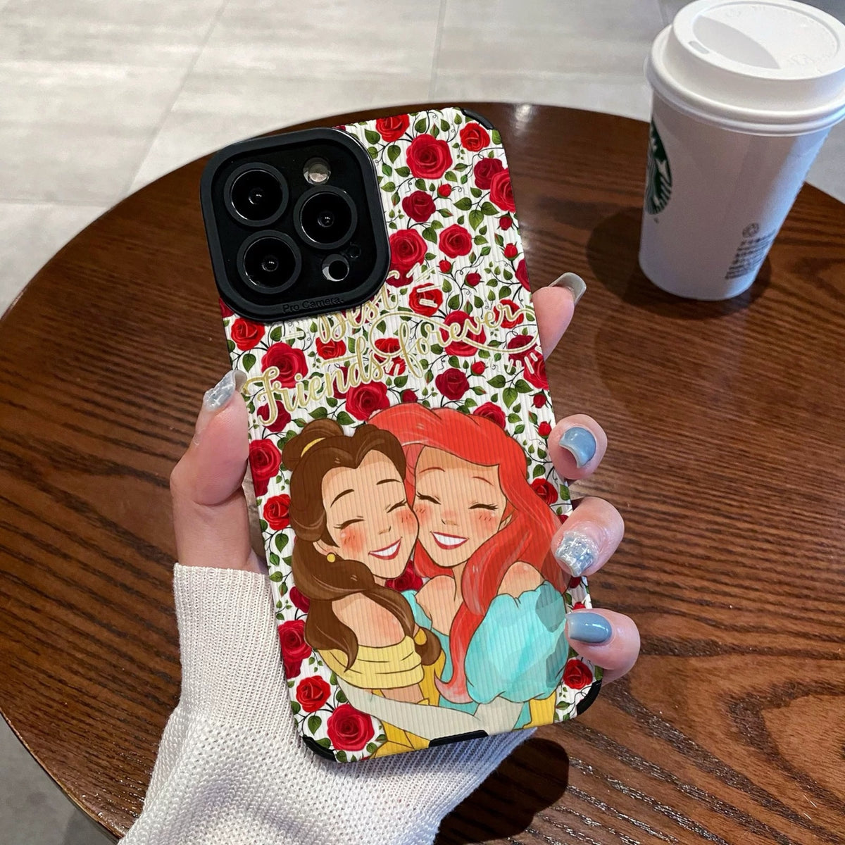 iPhone case with cartoon characters Princess Ariel and Belle on a wooden table with a Starbucks cup.