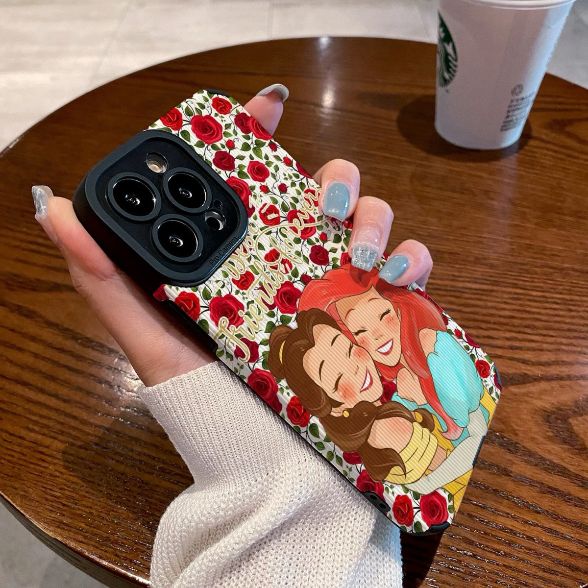 iPhone case with cartoon characters Princess Ariel and Belle on a wooden table with a Starbucks cup.
