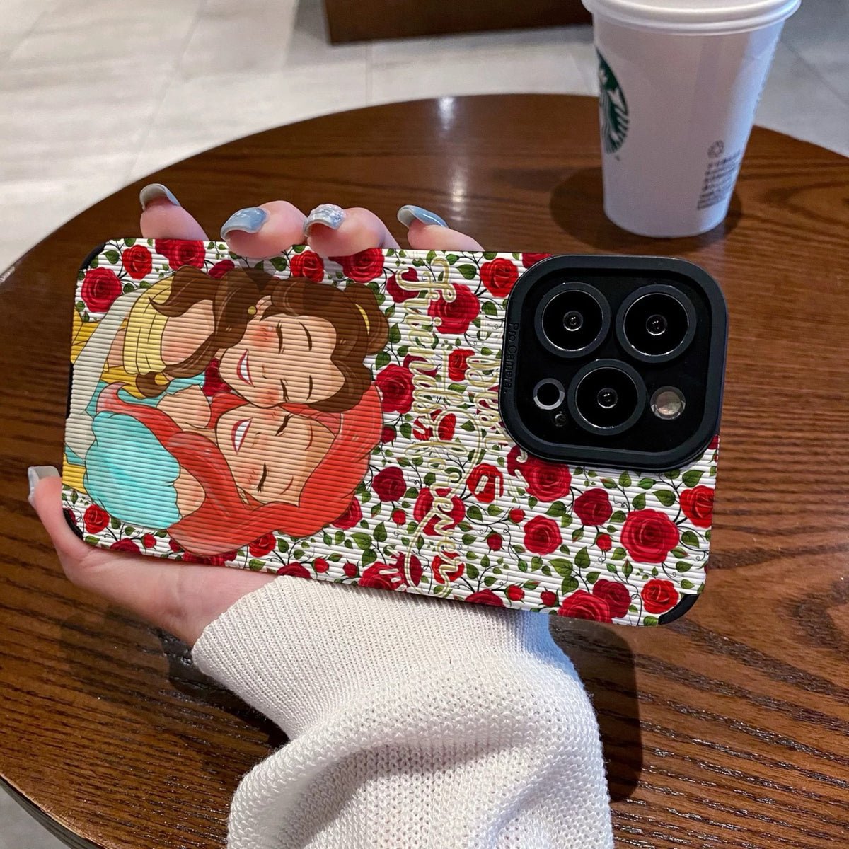 iPhone case with cartoon characters Princess Ariel and Belle on a wooden table with a Starbucks cup.