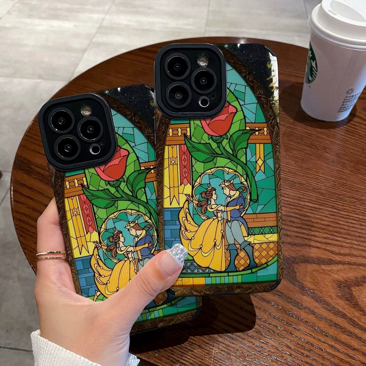iPhone case with stained glass design with belle and the prince held by a hand on a wooden table with a Starbucks cup.