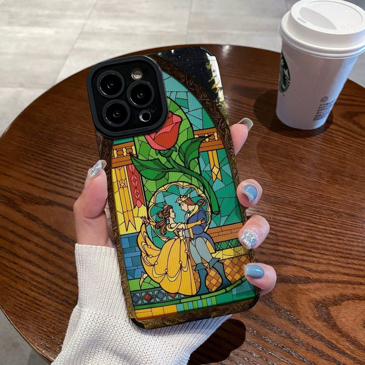 iPhone case with stained glass design with belle and the prince held by a hand on a wooden table with a Starbucks cup.