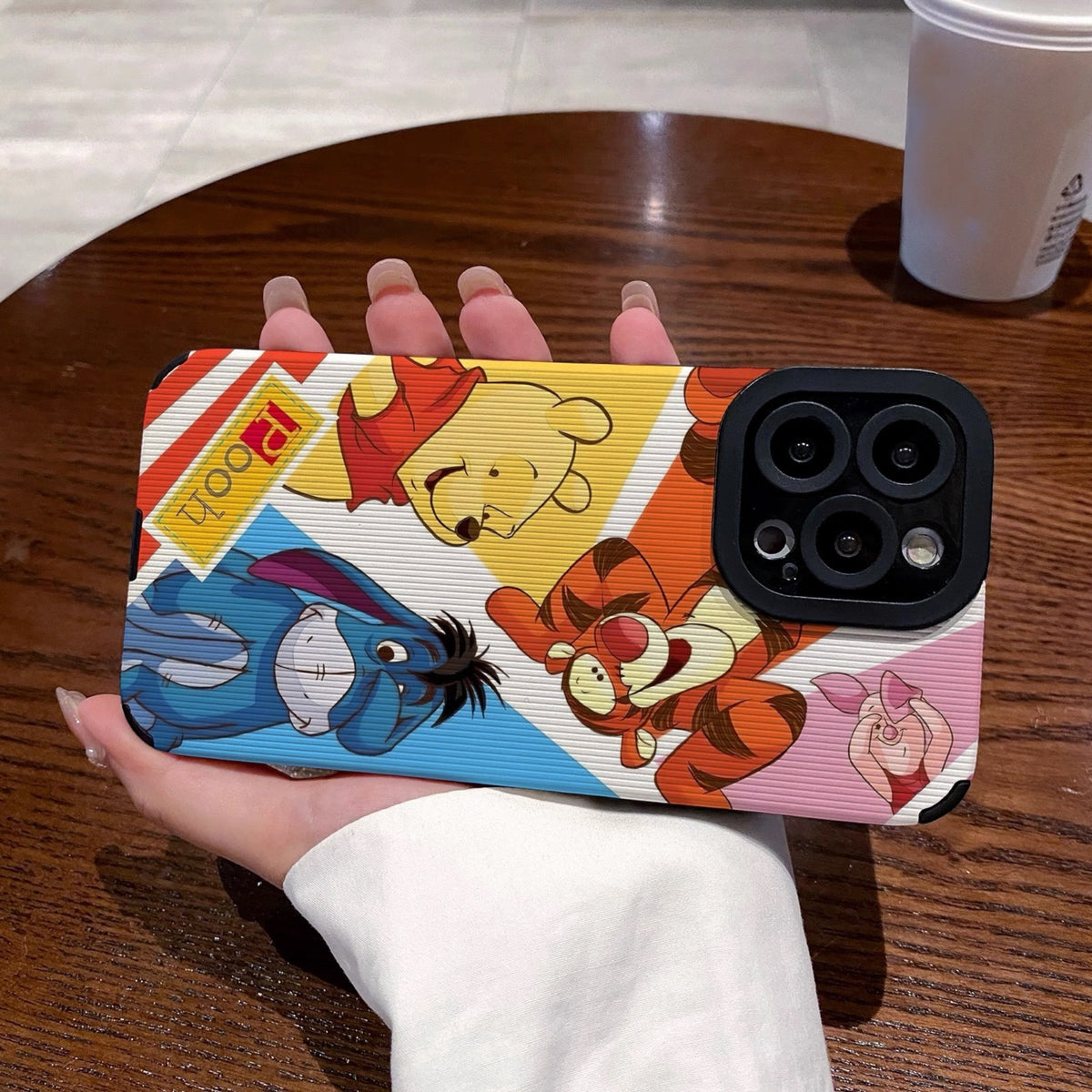 Phone case with cartoon characters winnie the pooh and friends held by a hand on a wooden table with a coffee cup.