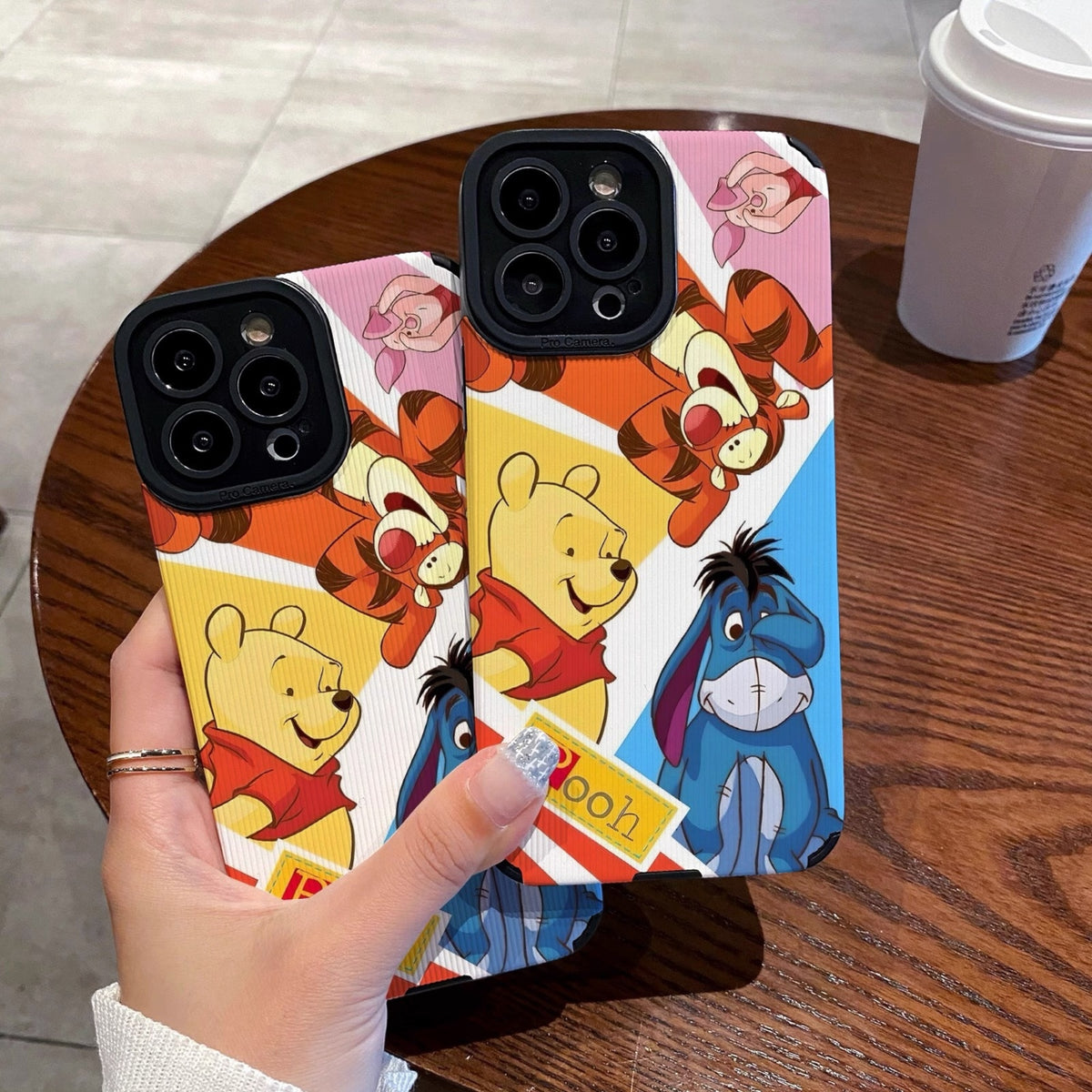 Phone case with cartoon characters winnie the pooh and friends held by a hand on a wooden table with a coffee cup.