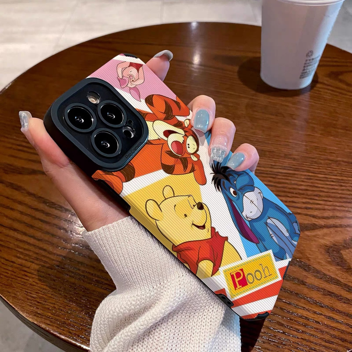 Phone case with cartoon characters winnie the pooh and friends held by a hand on a wooden table with a coffee cup.