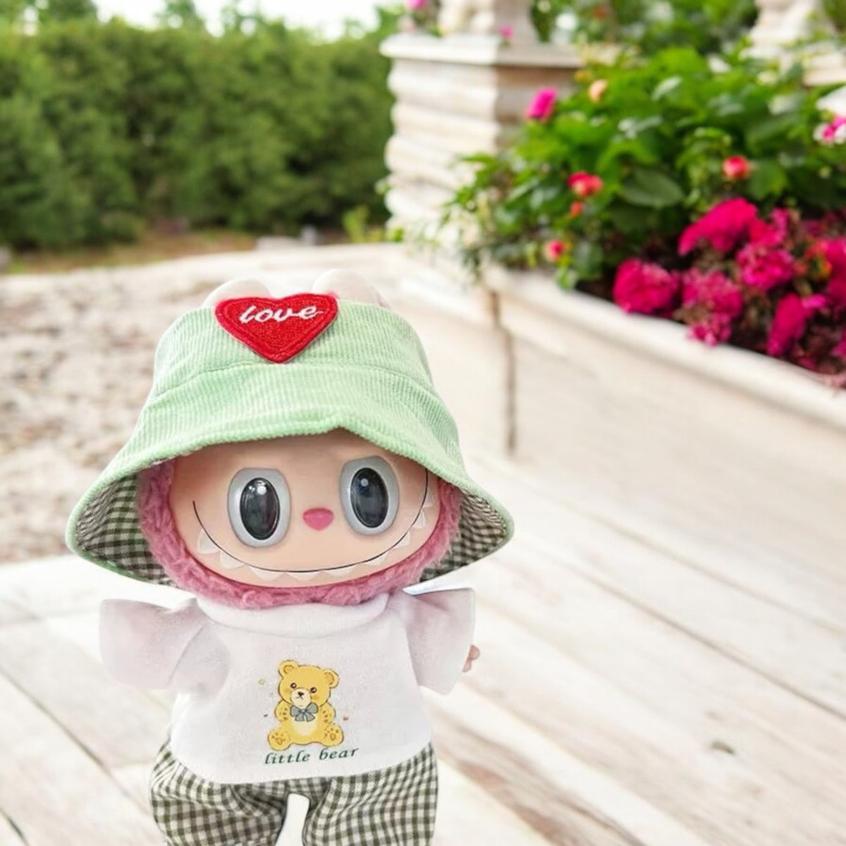 Stuffed toy with a green hat and pink face, sitting outdoors with flowers in the background.