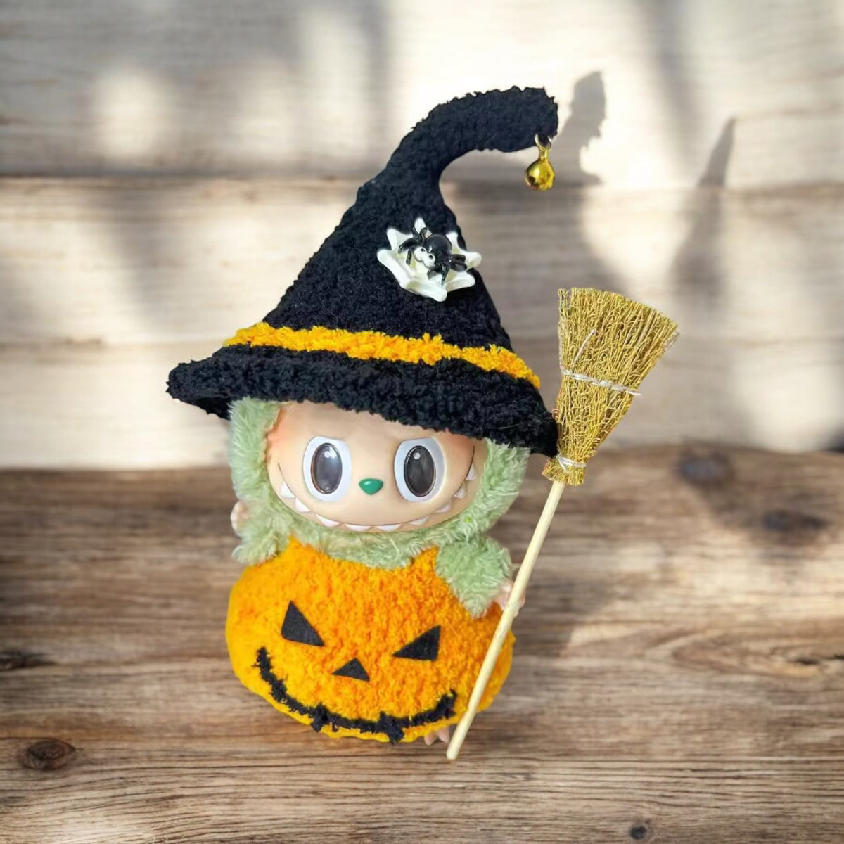 Halloween-themed plush toy with a witch hat, broom, and pumpkin costume on a wooden surface.