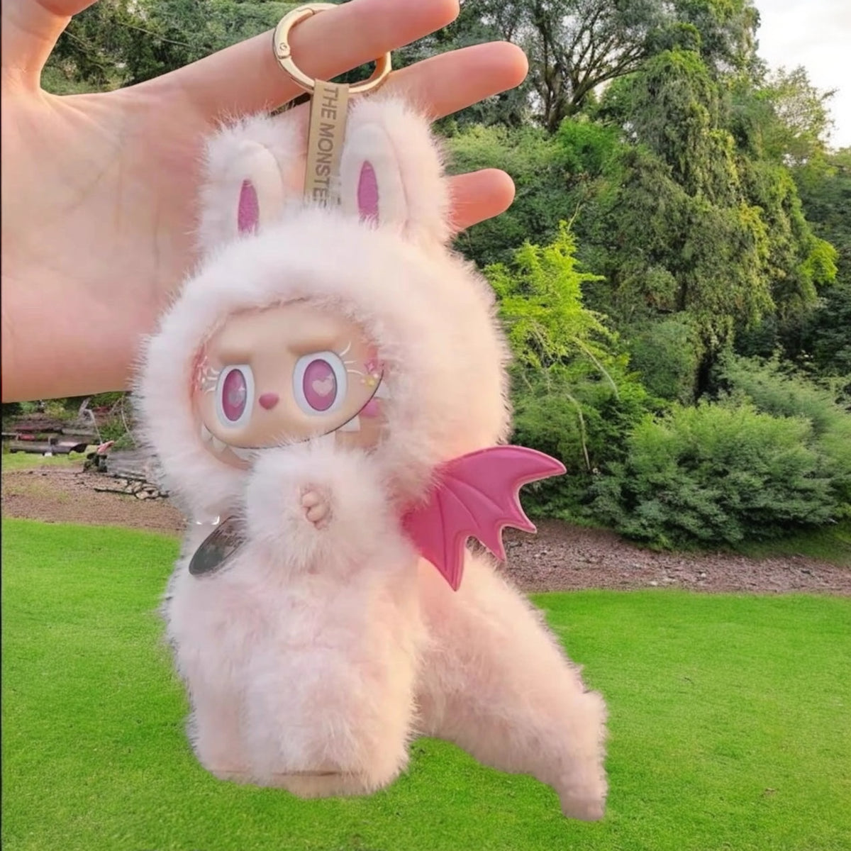 Furry plush keychain with pink zimomo dinosaurs outfit held by a hand outdoors.